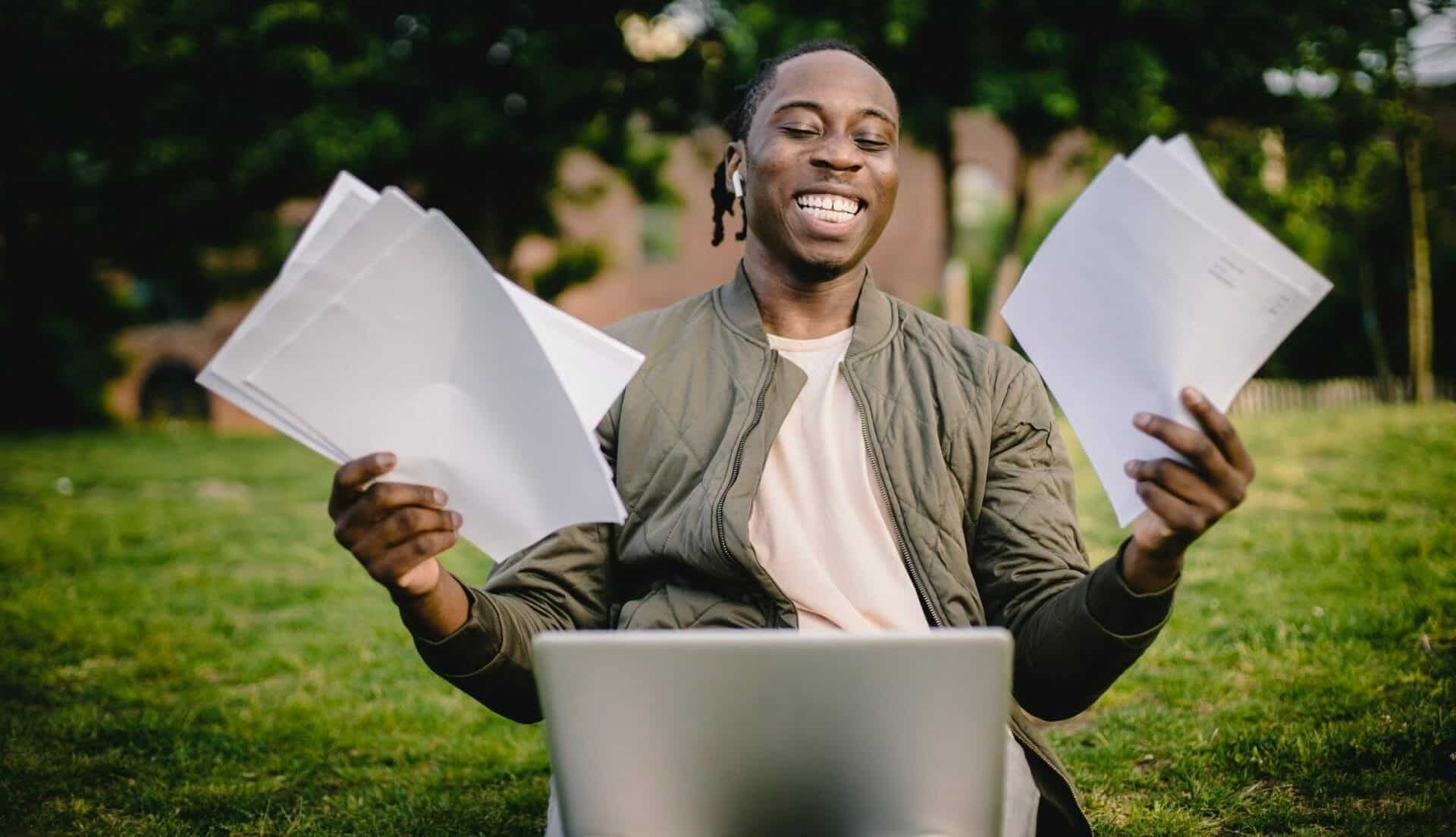 Man working on laptop outdoors, holding papers, looking like he's achieved something.